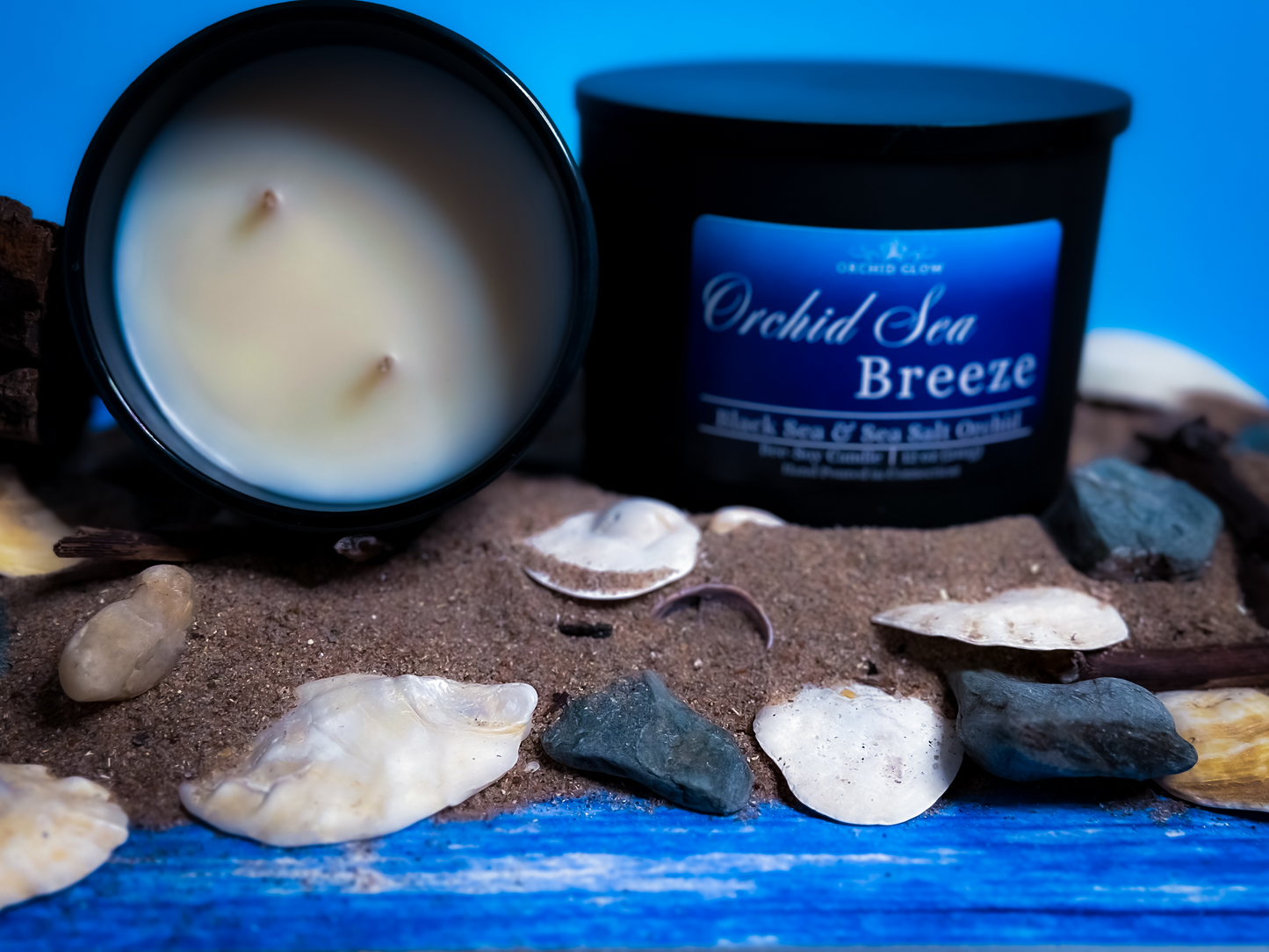 A black candle jar with a label reading 'Orchid Sea Breeze', placed on a bed of sand with shells and driftwood around it, against a blue background.
