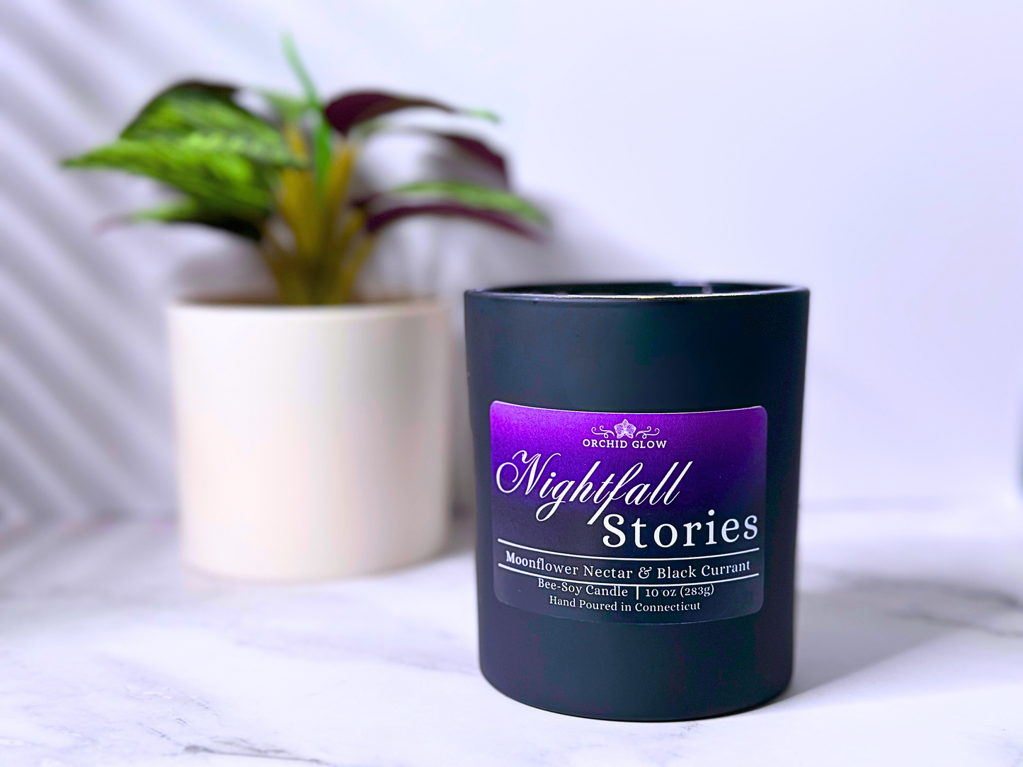 A black candle jar with a purple label that reads 'Nightfall Stories', with a white plant pot in the background.