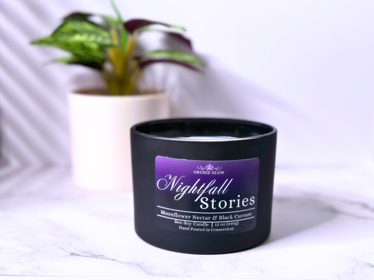 A black candle jar with a purple label that reads 'Nightfall Stories', placed on a white surface with a plant in the background.