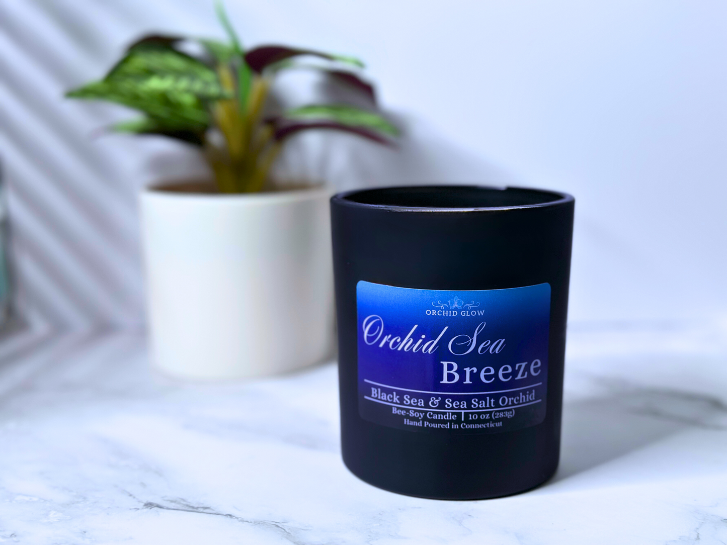 A black candle jar with a label reading 'Orchid Sea Breeze', placed on a marble surface and flower behind.