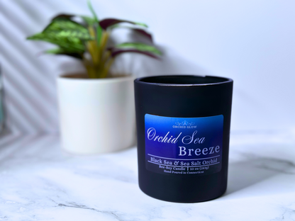 A black candle jar with a label reading 'Orchid Sea Breeze', placed on a marble surface and flower behind.